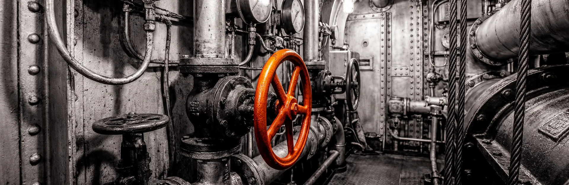 Detail eines Maschinenraums in Schwarzweiß mit orangefarbenem Ventilrad.